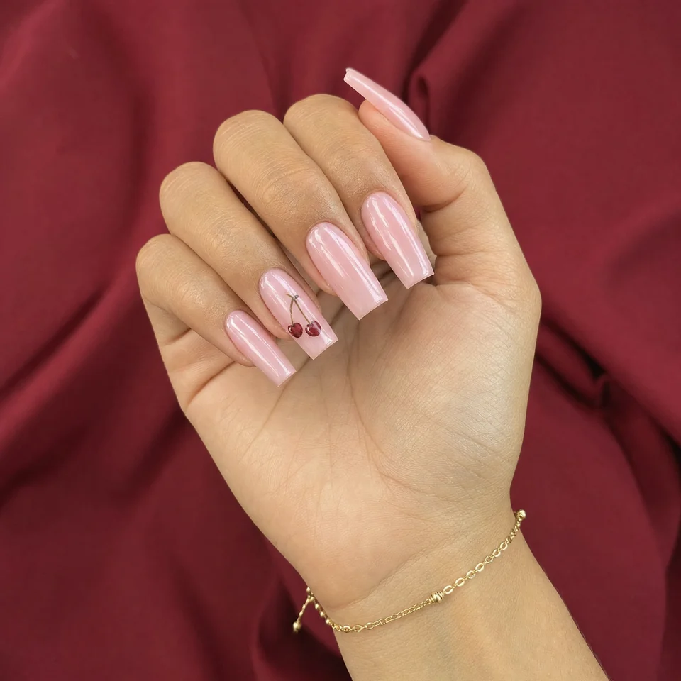 A gently curled hand with long pink square nails, a small cherry nail design and a gold bracelet against burgundy fabric.